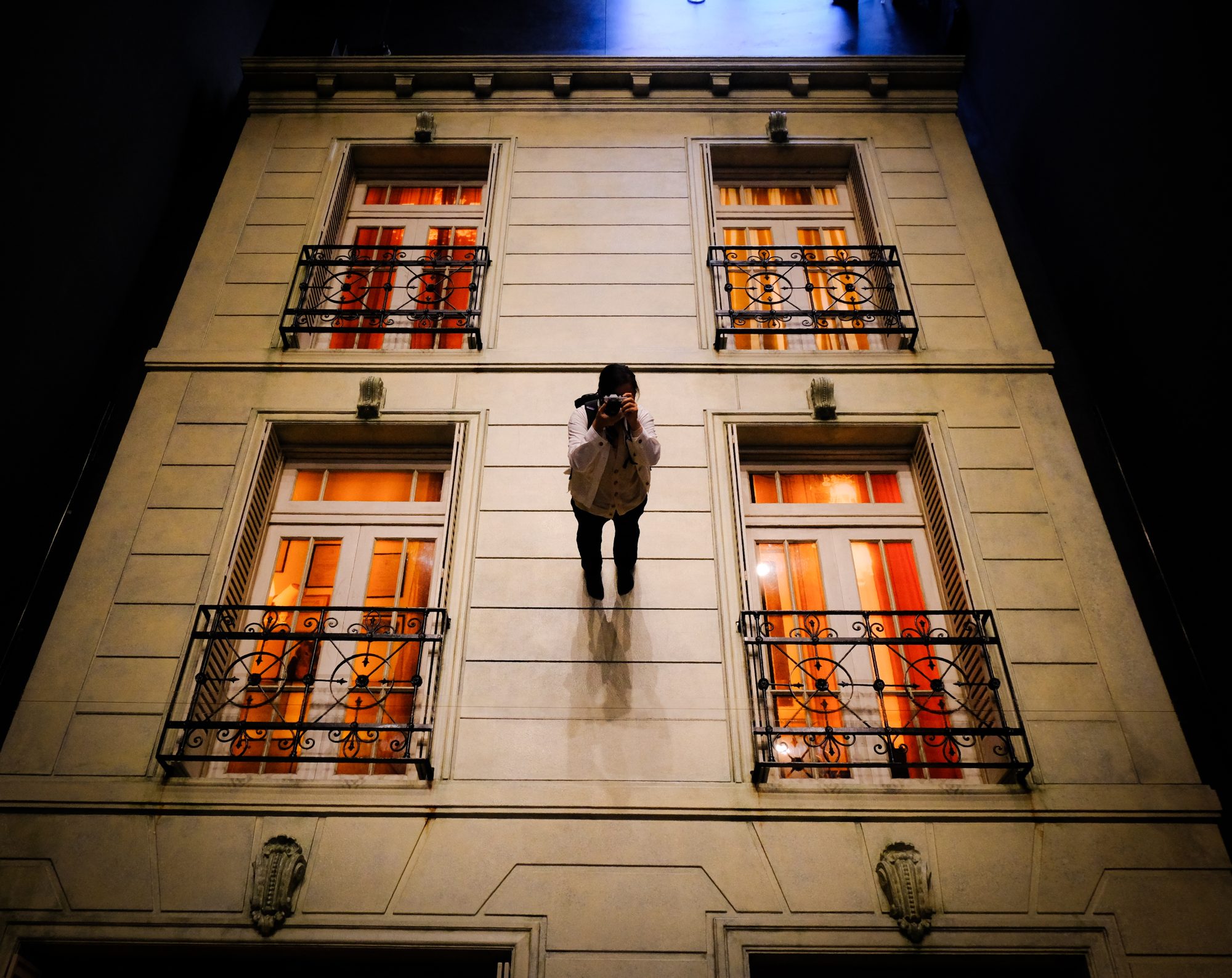 A woman holding a camera towards the mirror, but it looks like she is standing on the wall of an architecture. Trick view